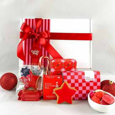 A Christmas-themed gift hamper featuring a white box wrapped in red and white striped paper with a large red ribbon, surrounded by festive treats including red star-shaped cookies, a red-and-white checkered box, candy canes, and red ornaments on a white background.