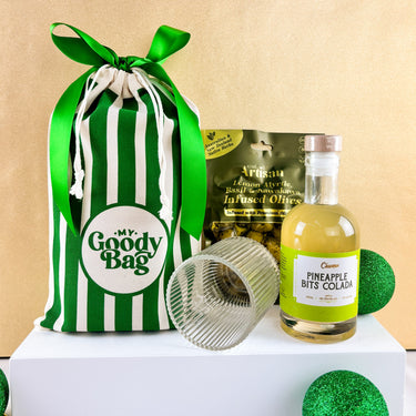 pineapple bits colada alcoholic drink with reeded glass and kiwi artisan olives flank green striped goody bag against gold background by My Goody Bag