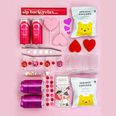 Gifting set with drinks, snacks, and decorative items on a pink background