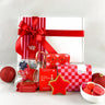 A Christmas-themed gift hamper featuring a white box wrapped in red and white striped paper with a large red ribbon, surrounded by festive treats including red star-shaped cookies, a red-and-white checkered box, candy canes, and red ornaments on a white background.