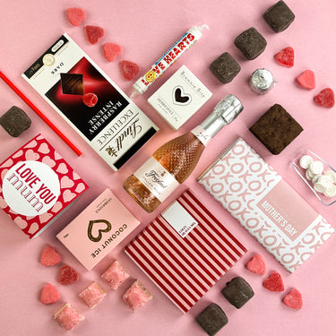 Gourmet gift set with chocolates, cookies, and a bottle of wine on a pink background.