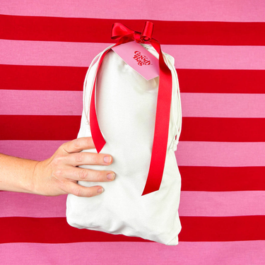hand held natural canvas drawstring bag with red ribbon and My Goody Bag tag against red and prink striped background